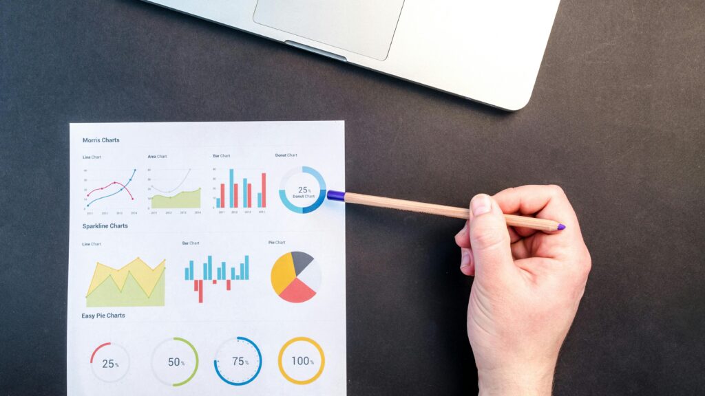 Hand holding pencil reviewing colorful data charts on desk with laptop.
